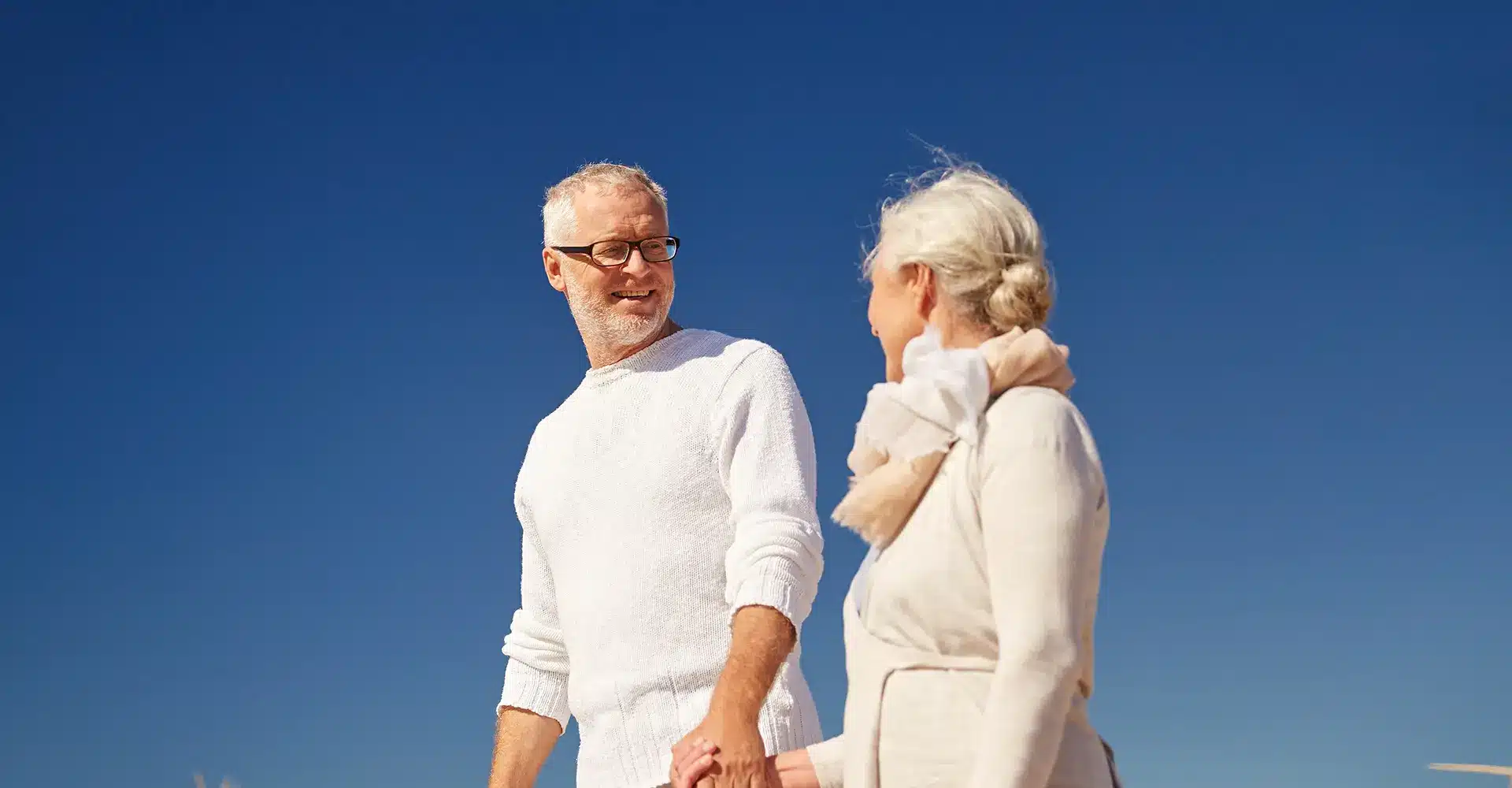 an elderly couple both with white hair walk hand in hand outdoors against the backdrop of a clear blue sky the man wears glasses and a white sweater and the woman who benefits from hormone therapy dons a light beige coat and a light pink scarf both smile at each other
