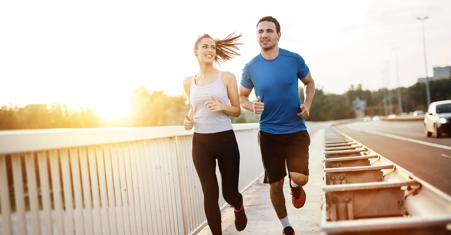 a woman in a gray tank top and black leggings runs alongside a man in a blue t shirt and black shorts on a bridge during sunset both are smiling and appear to be enjoying their run perhaps fueled by the energy boost of iv therapy the background includes trees and a clear sky