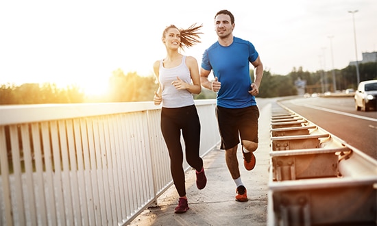 a man and a woman are jogging side by side on a bridge during sunset embodying wellness the man is wearing a blue t shirt and black shorts while the woman sports a white tank top and black leggings they appear to be enjoying their run with the sun setting in the background