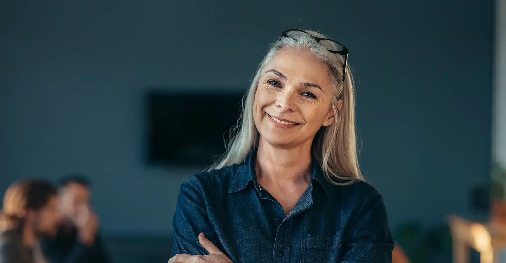 a smiling woman with long gray hair wearing a dark denim shirt and glasses perched on her head stands with her arms crossed the background is blurred showing two indistinct people sitting and discussing cosmetic surgery
