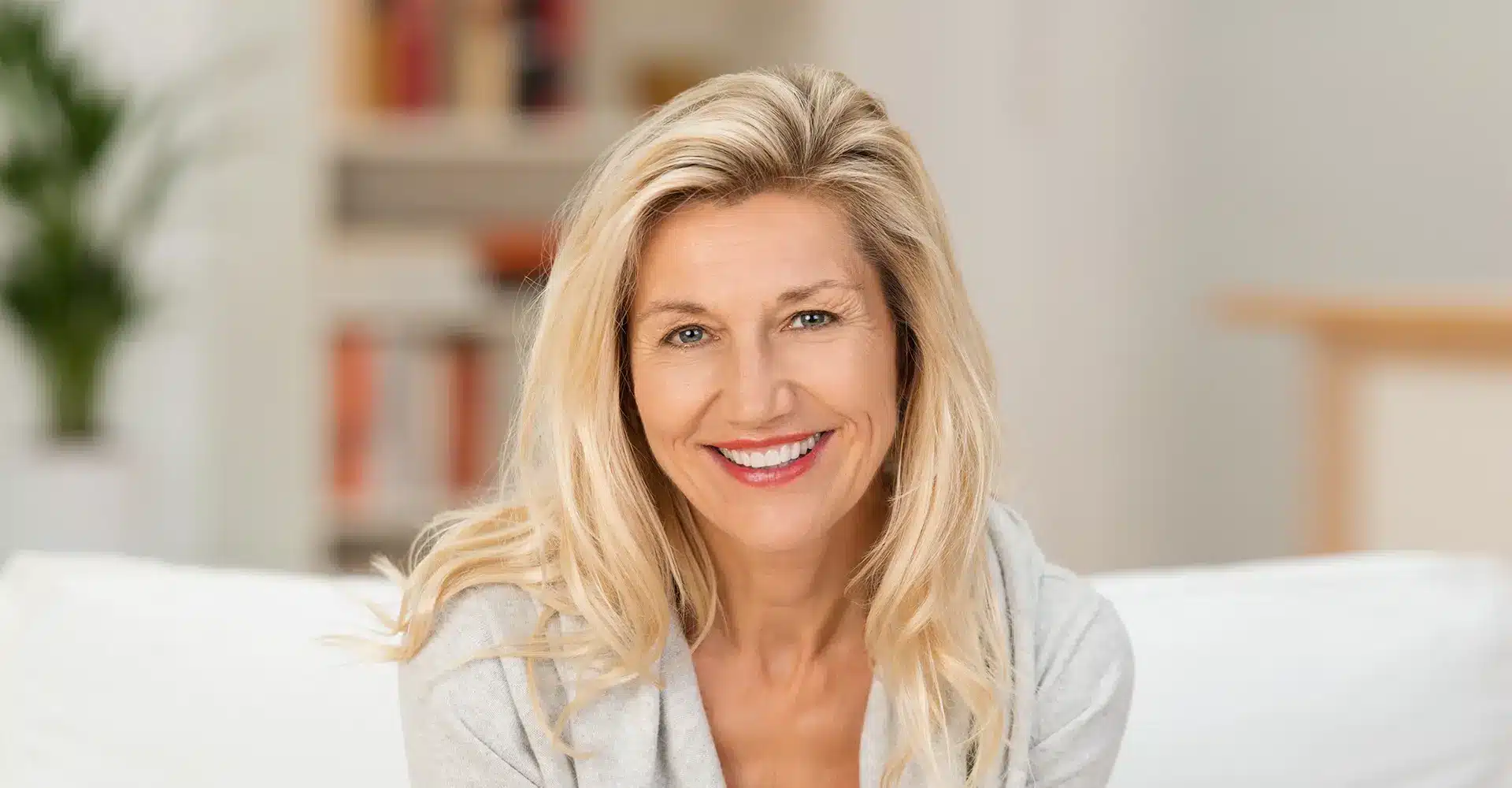 a smiling woman with long blonde hair is sitting indoors on a white couch she is wearing a light gray sweater looking radiant after recent cosmetic treatments the blurred background includes bookshelves and a green plant making the overall scene appear bright and welcoming