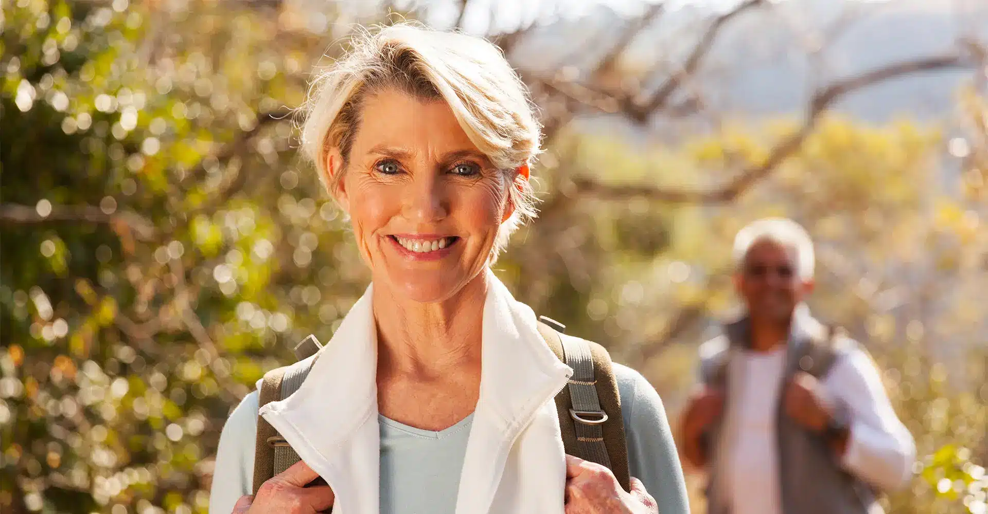 a smiling woman with short gray hair stands outdoors wearing a light jacket and backpack in the blurred background another individual also with gray hair can be seen wearing a backpack they appear to be on a hiking trail surrounded by trees enjoying nature and promoting their skin health