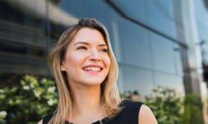 a woman with shoulder length blonde hair is smiling and looking away from the camera she is wearing a black top and is standing in front of a modern glass building with greenery in the background the sky is clear and the overall atmosphere is bright and cheerful highlighting her natural beauty without any hint of cosmetic surgery