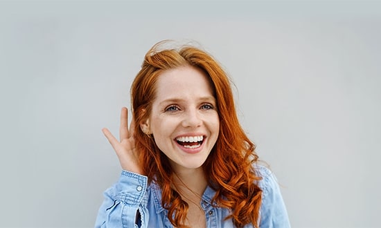 a red haired person wearing a light blue shirt smiles brightly against a plain background raising one hand near their ear in a listening gesture as though intrigued by the latest advancements in cosmetic surgery