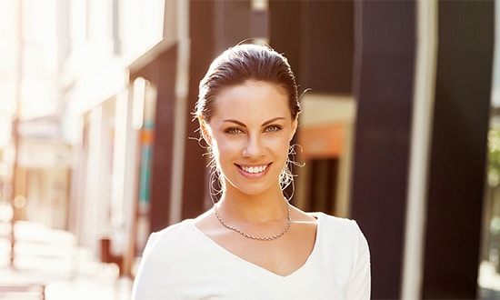 a woman with brown hair pulled back is smiling warmly at the camera she is wearing a white top and a delicate necklace the background an urban setting with blurred buildings and sunlight streaming in gives a bright cheerful ambiance subtly hinting at the confidence often gained through cosmetic surgery